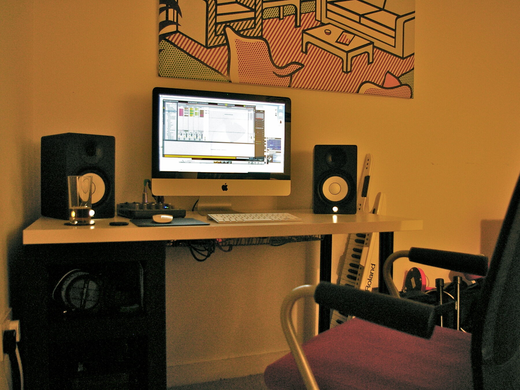 An iMac and monitor speakers on a white desk against a mustard wall with large artwork